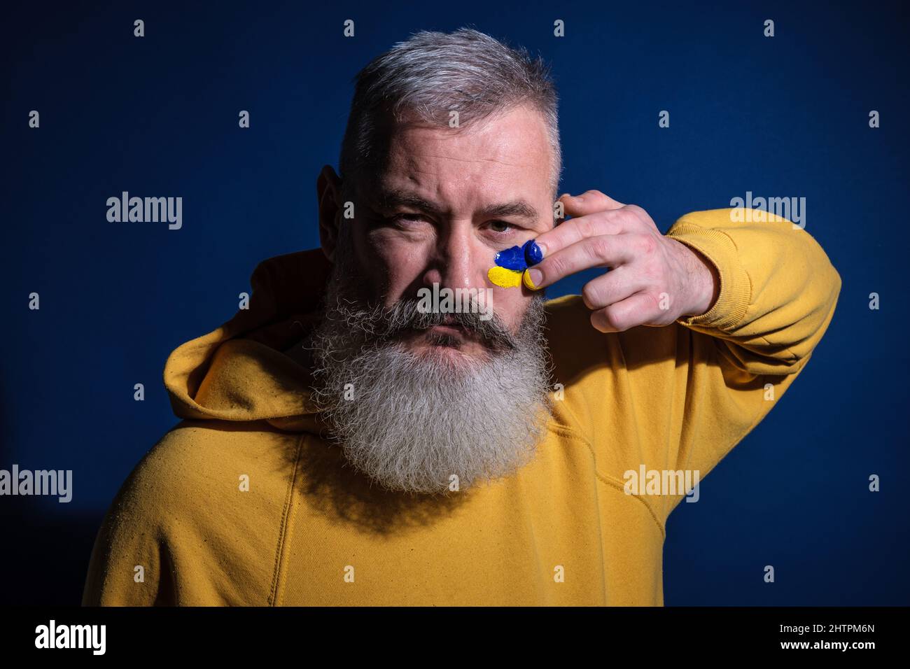Mature bearded man dressed yellow hoodie draws Ukrainian flag on his face, protest against war Stock Photo