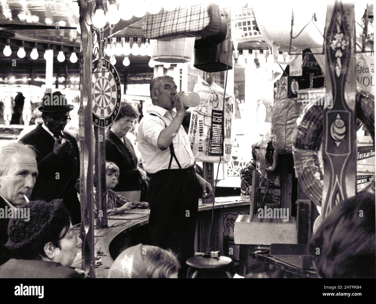 A stall holder takes a welcome drink at his typical fairground  prize bingo stall at Manningham,  Bradford, West Yorkshire, England, sometime in the early 1960's . Various prizes such as a bathroom set and dartboard can be see hanging on the circular stall supports. Stock Photo