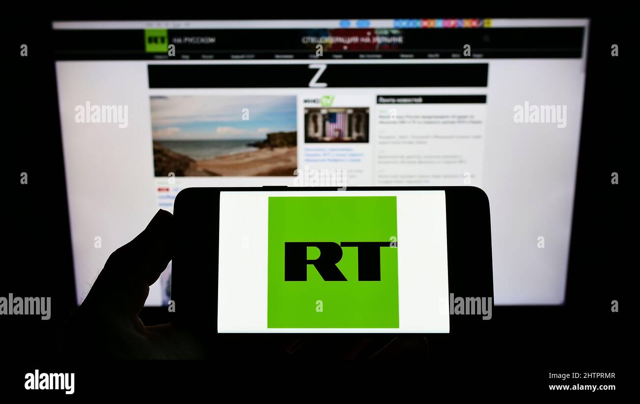 Person holding smartphone with logo of Russian state-controlled television company RT on screen in front of website. Focus on phone display. Stock Photo