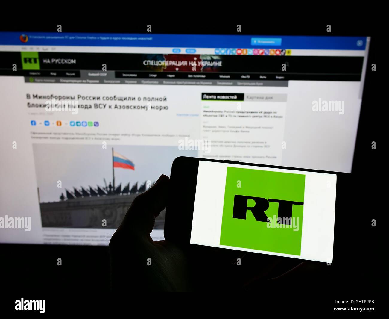 Person holding mobile phone with logo of Russian state-controlled television company RT on screen in front of web page. Focus on phone display. Stock Photo