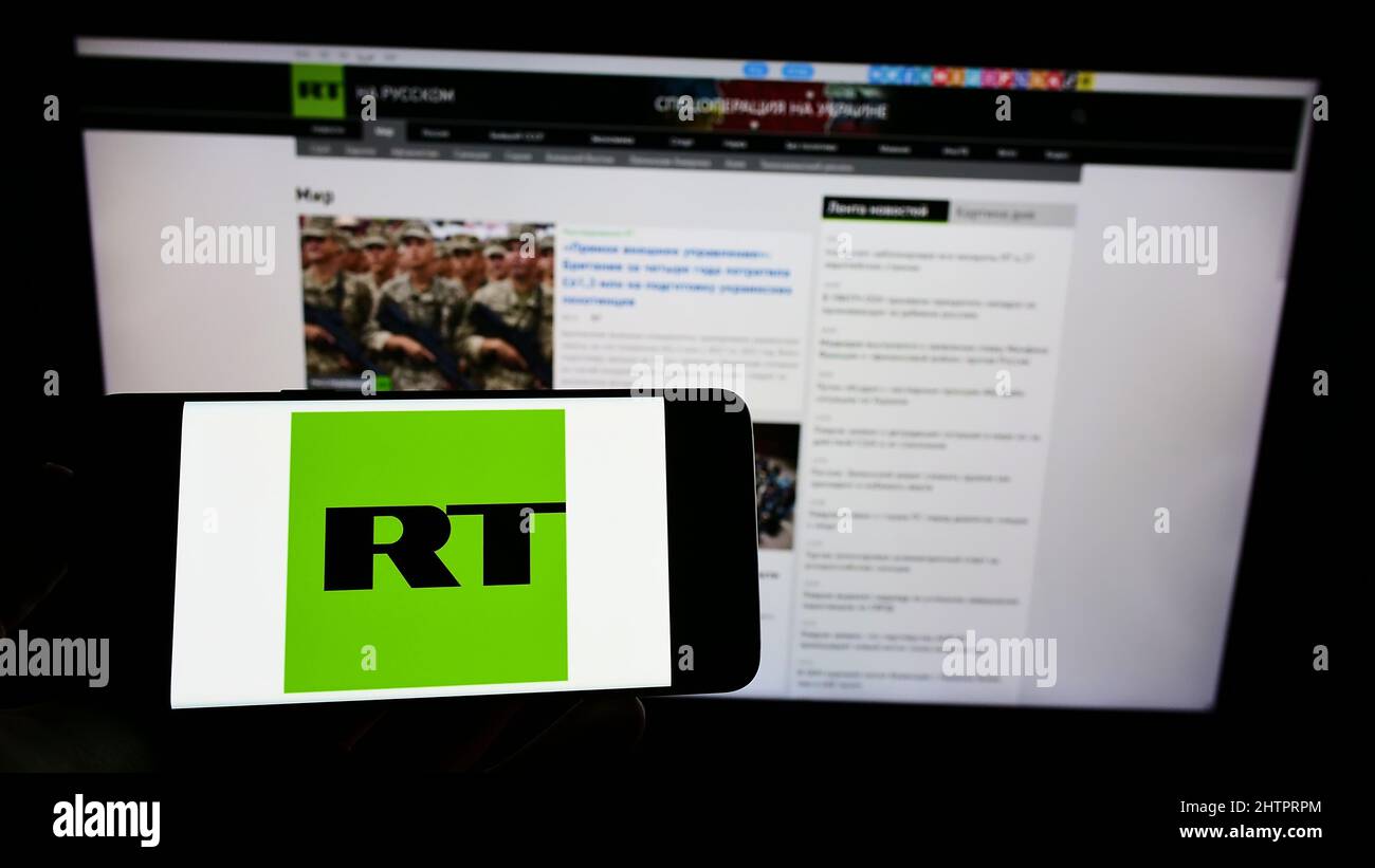 Person holding cellphone with logo of Russian state-controlled television company RT on screen in front of webpage. Focus on phone display. Stock Photo