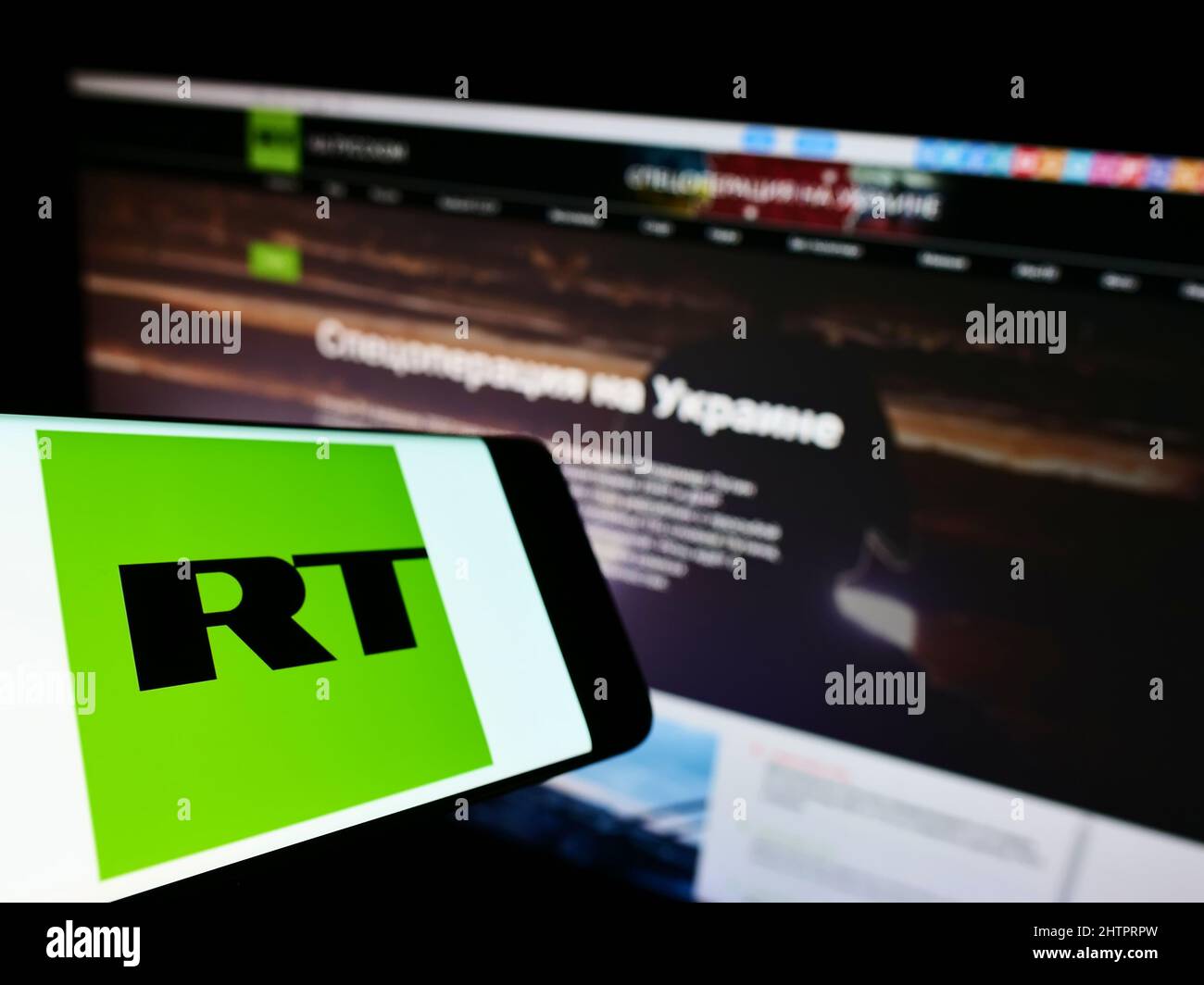 Mobile phone with logo of Russian state-controlled television company RT on screen in front of website. Focus on center-left of phone display. Stock Photo