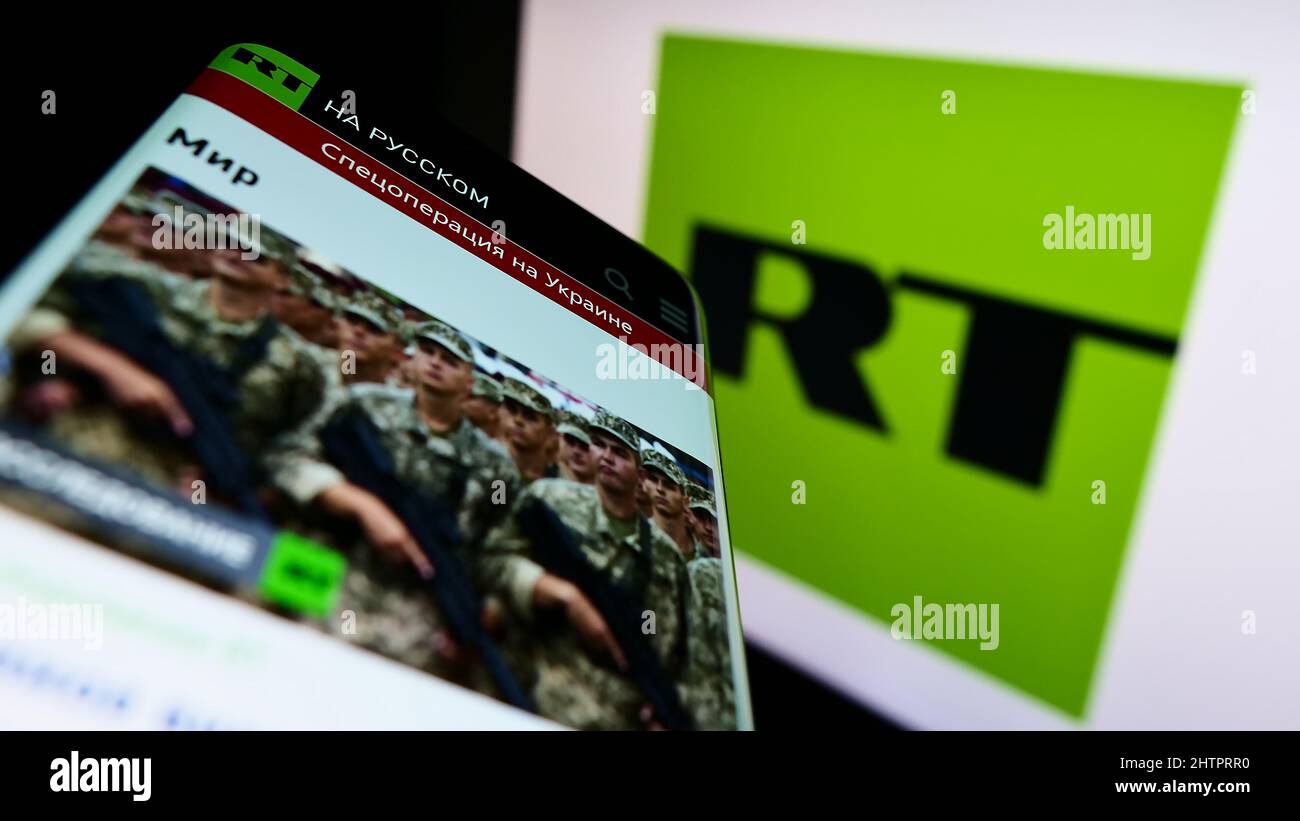 Smartphone with website of Russian state-controlled television company RT on screen in front of business logo. Focus on top-left of phone display. Stock Photo