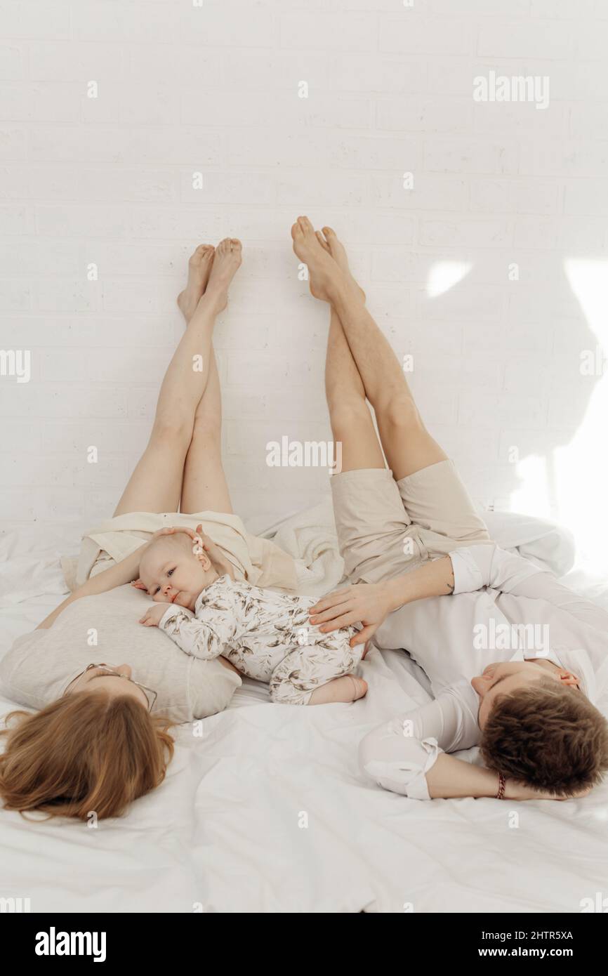 Young couple in white clothes lying with legs raised straight up high on white bed, caressing cute plump blue-eyed baby. Stock Photo