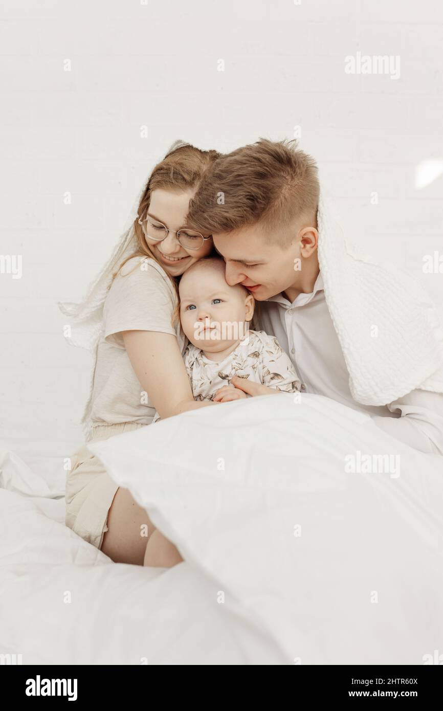 Portrait of young happy smiling beautiful family with cute cherubic infant baby in white clothes sitting on white bed. Stock Photo