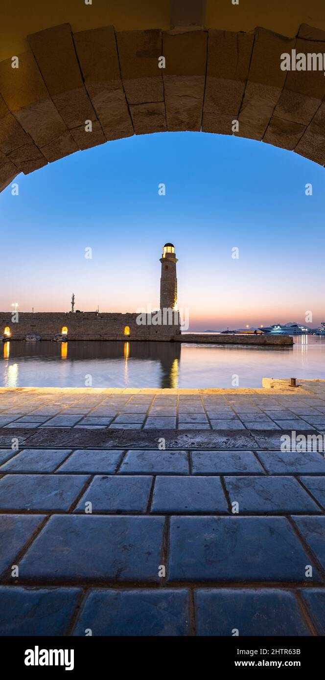Illuminated lighthouse at dusk view through a medieval arch in the old Venetian harbour, Rethymno, Crete, Greece Stock Photo