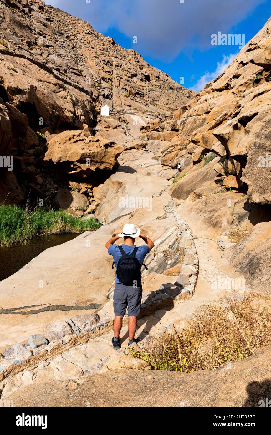 Hiker with backpack looking at the old chapel among rock canyons, Barranco de las Penitas, Fuerteventura, Canary Islands, Spain Stock Photo