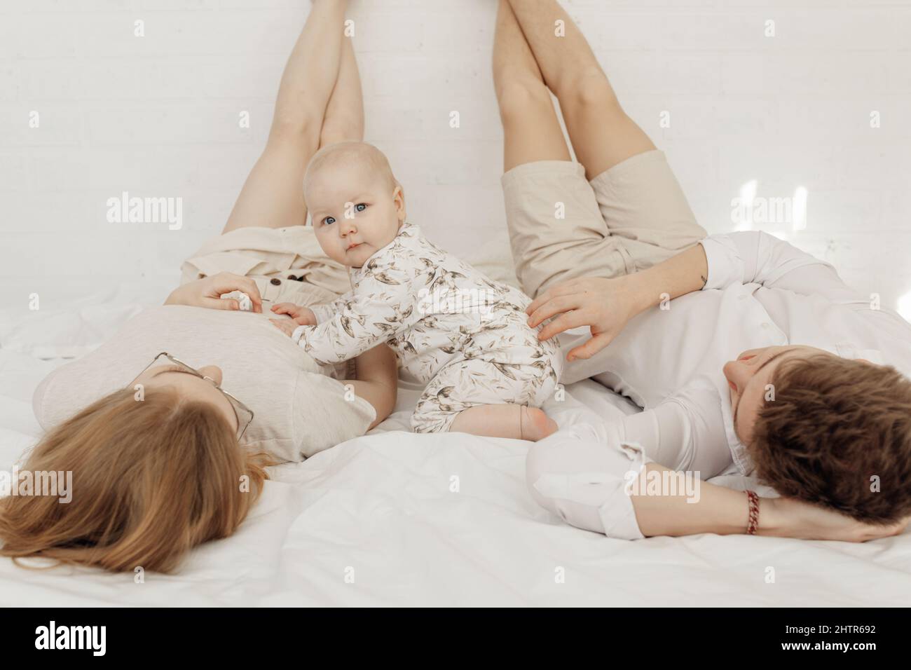Young couple in white clothes lying with legs raised straight up high on white bed with awesome chubby blue-eyed baby. Stock Photo