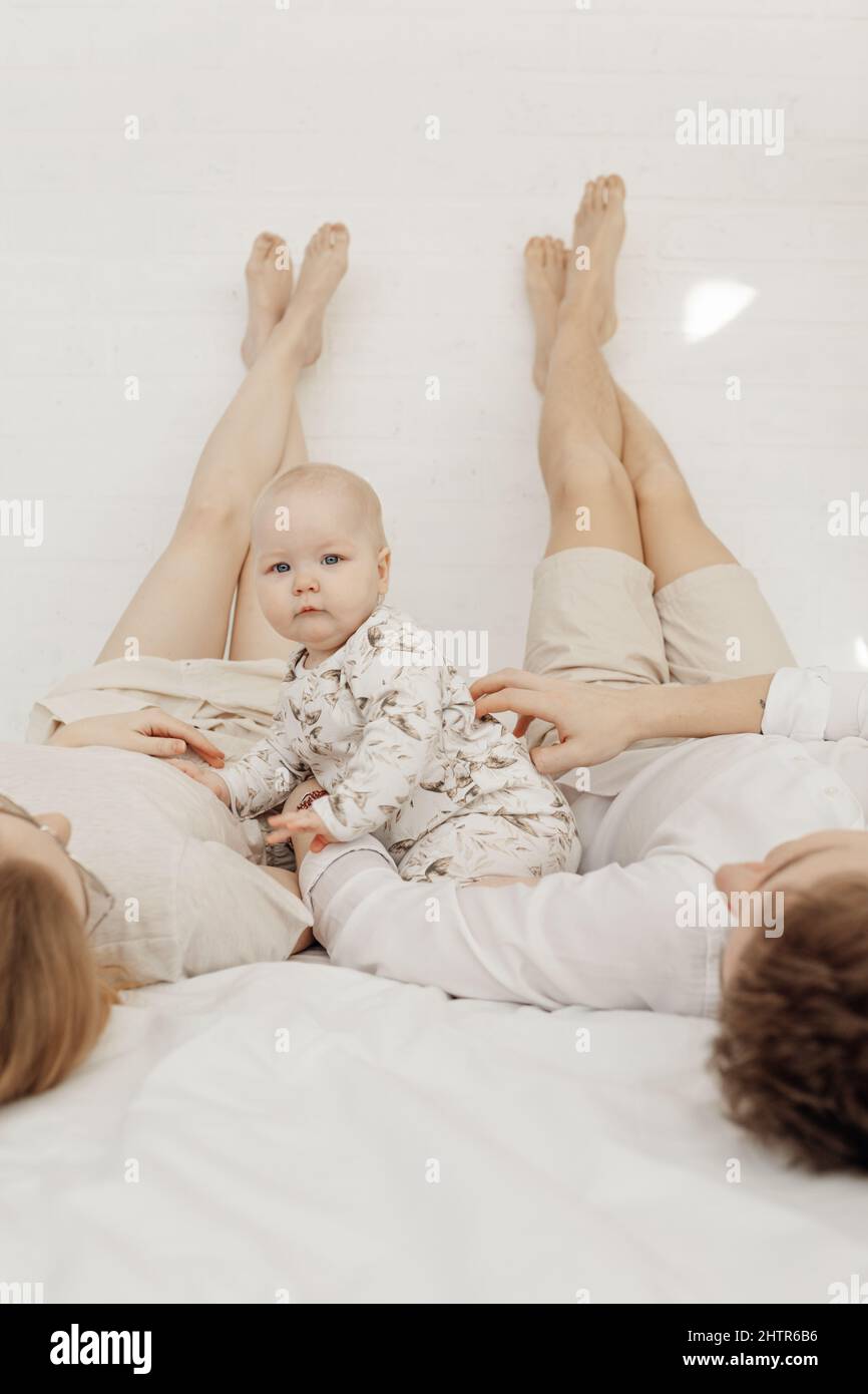 Young couple in white clothes lying with legs raised straight up high on bed with baby infant sitting between parents. Stock Photo