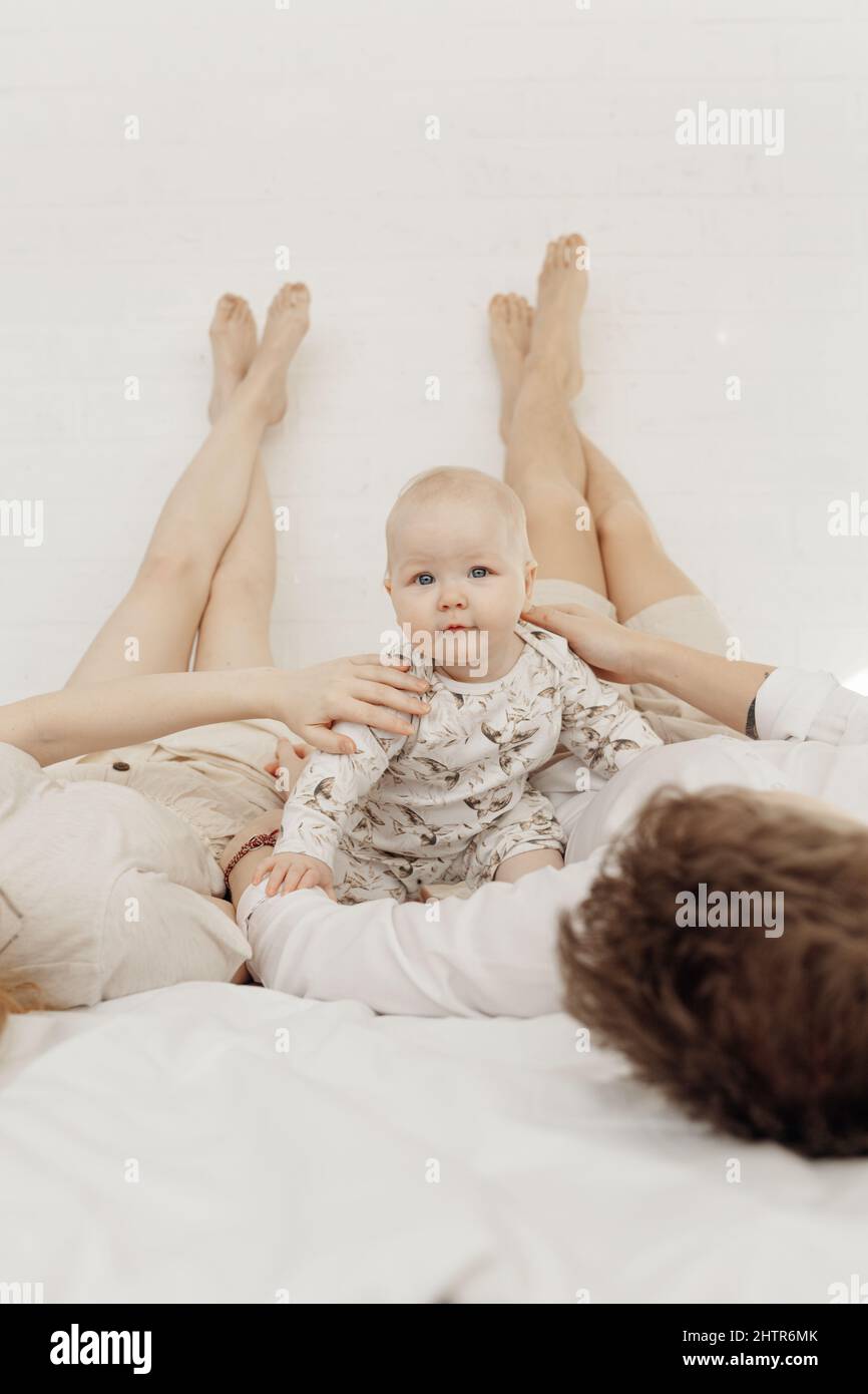 Young couple in white clothes lying with legs raised straight up high on bed putting hands on shoulders of plump baby. Stock Photo