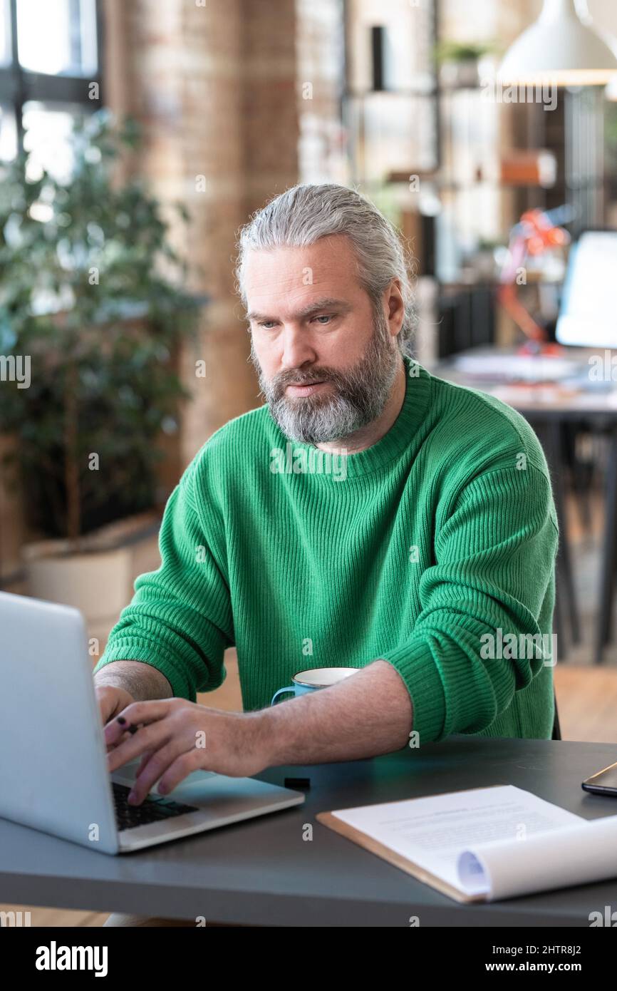 Mature bearded businessman sitting on his workplace and typing on laptop during work at office Stock Photo