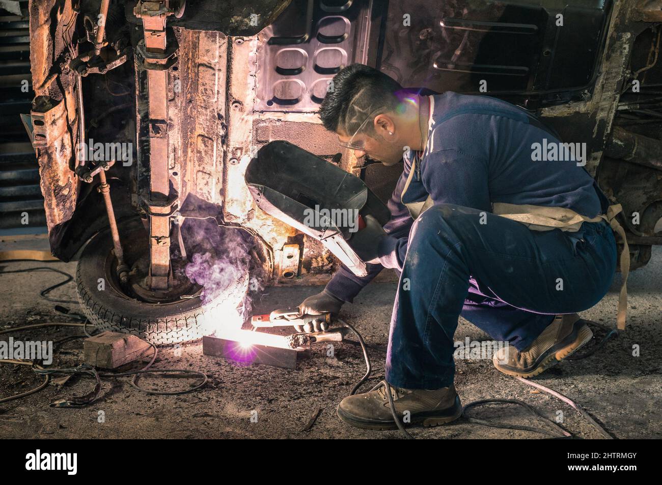 Young man mechanic worker repairing old vintage car body in messy garage - Safety at work and protection wear - Guy with cool hair cut at vehicle reno Stock Photo