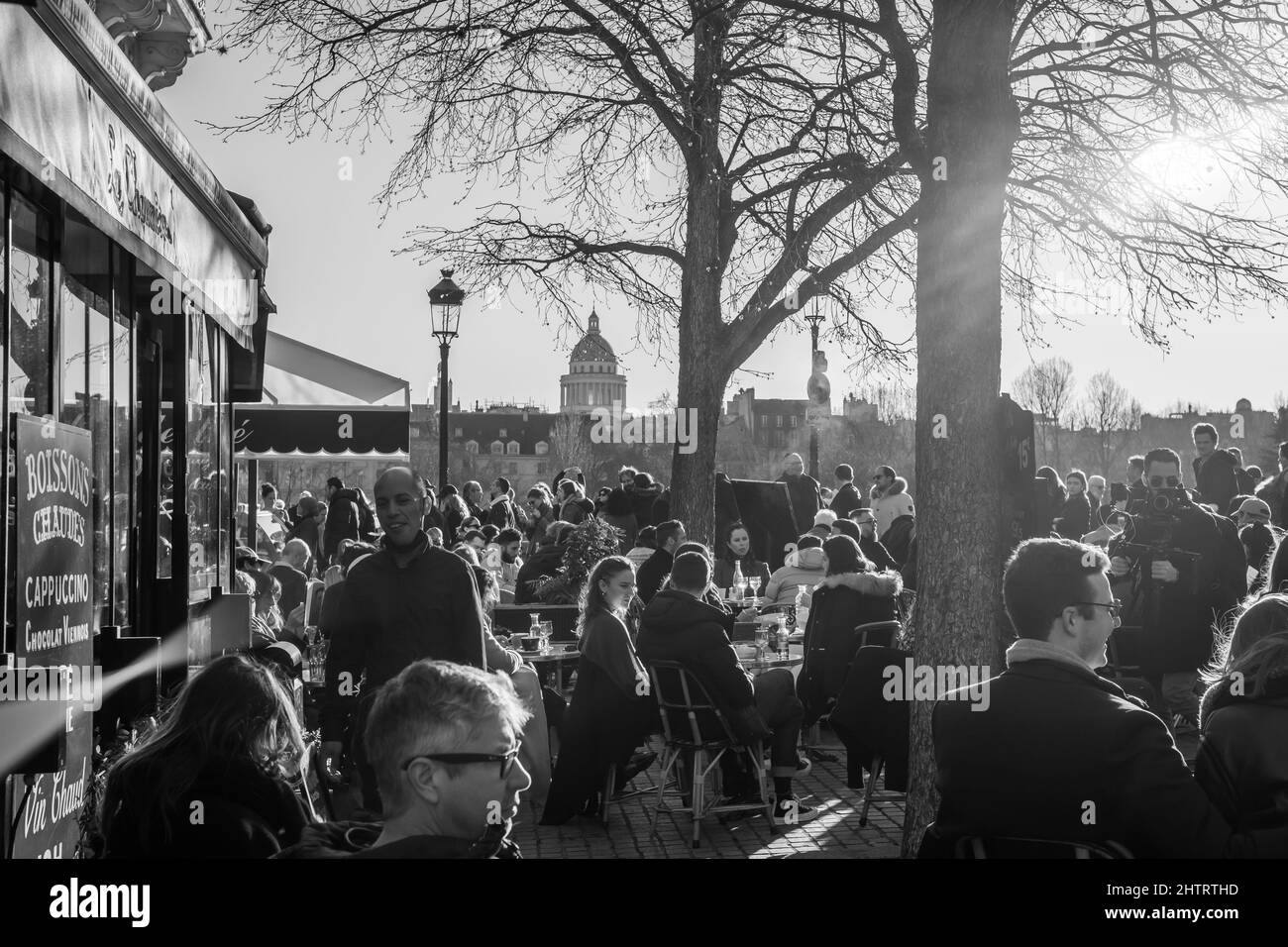 Paris, France - February 27, 2022 : Tourists and Parisians enjoying food and drinks at a typical French Café Restaurant in Paris on a sunny day in black and white Stock Photo