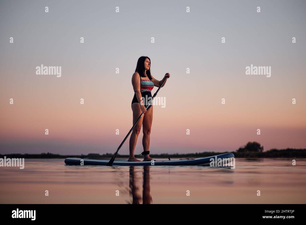 Sup boarding woman of middle age rowing with puddle looking away on rippled lake with astonishing pink sky in background wearing swimsuit in evening Stock Photo