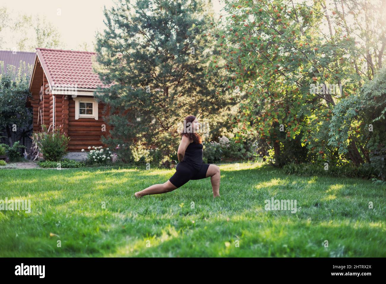 Blissful fat woman doing exercises for stretching legs green grass on backyard of cottage with wooden house and trees in background. Body positivity Stock Photo
