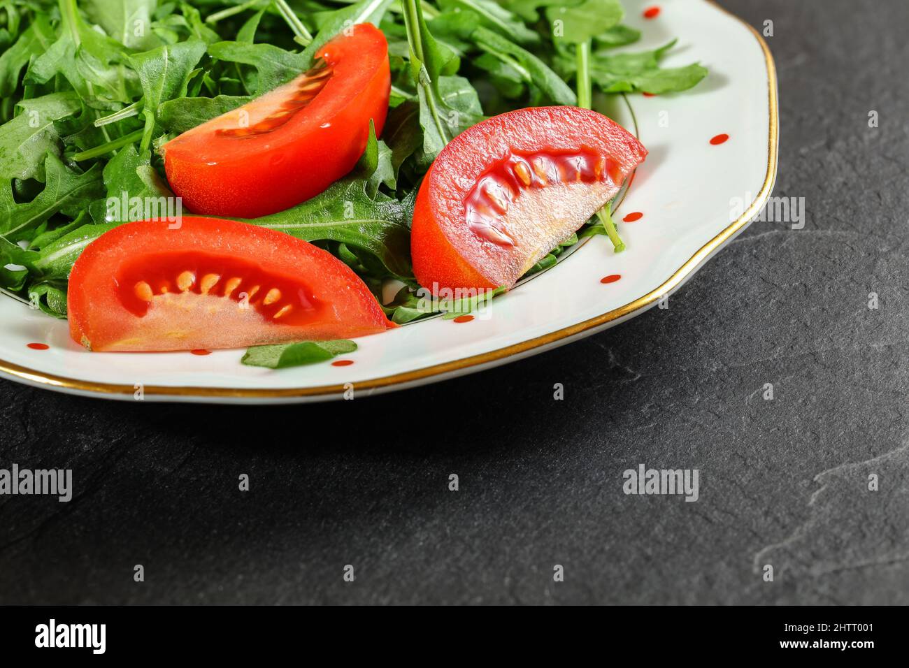 Plate with green leaves salad - arugula rocket - with fresh red tomatos on black stone desk, closeup detail Stock Photo