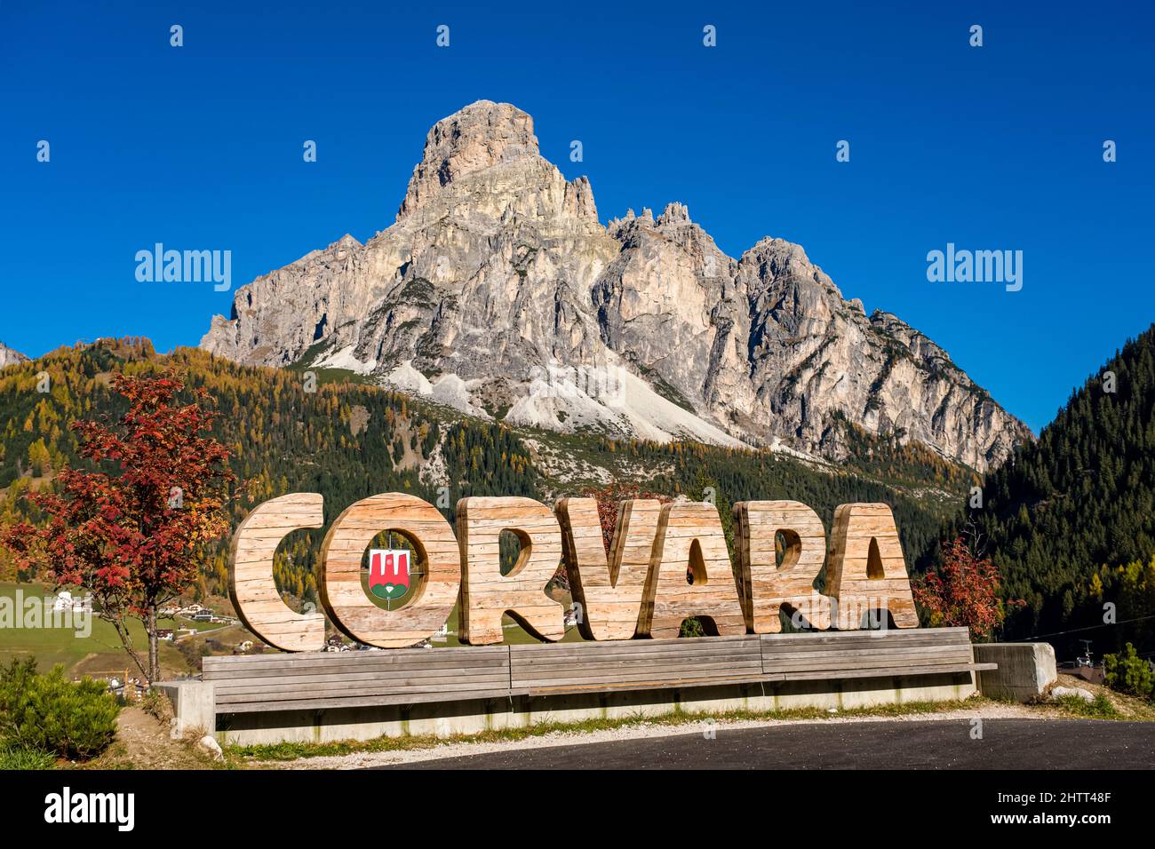Mountain Mt. Sassongher, Sass Songher, raising above the town and colorful trees in autumn, the name Corvara cut in wood. Stock Photo
