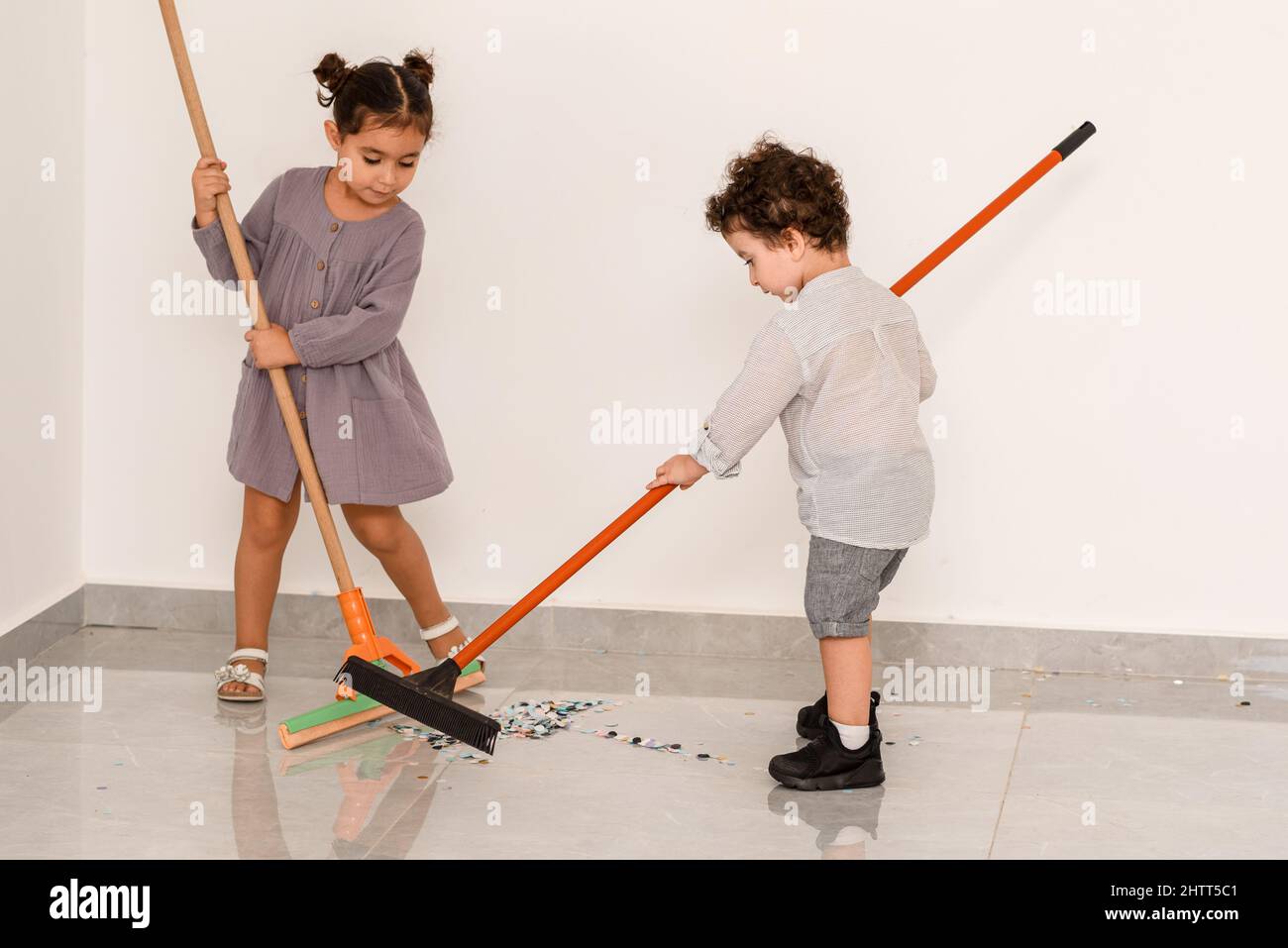 After party. Little curls boy and beautiful girl sweeping confetti with brooms. Brother and sister cleaning floor in a home with mops. Children cleaning the living room together. Stock Photo