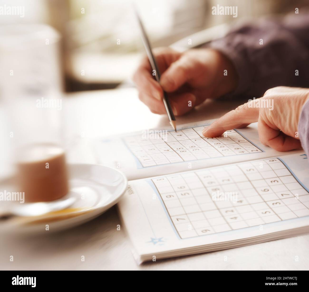 Woman trying to solve Sudoku Puzzle - window, sunlight, and coffee. Woman trying to solve Sudoku Puzzle - window, sunlight, and coffee. Stock Photo