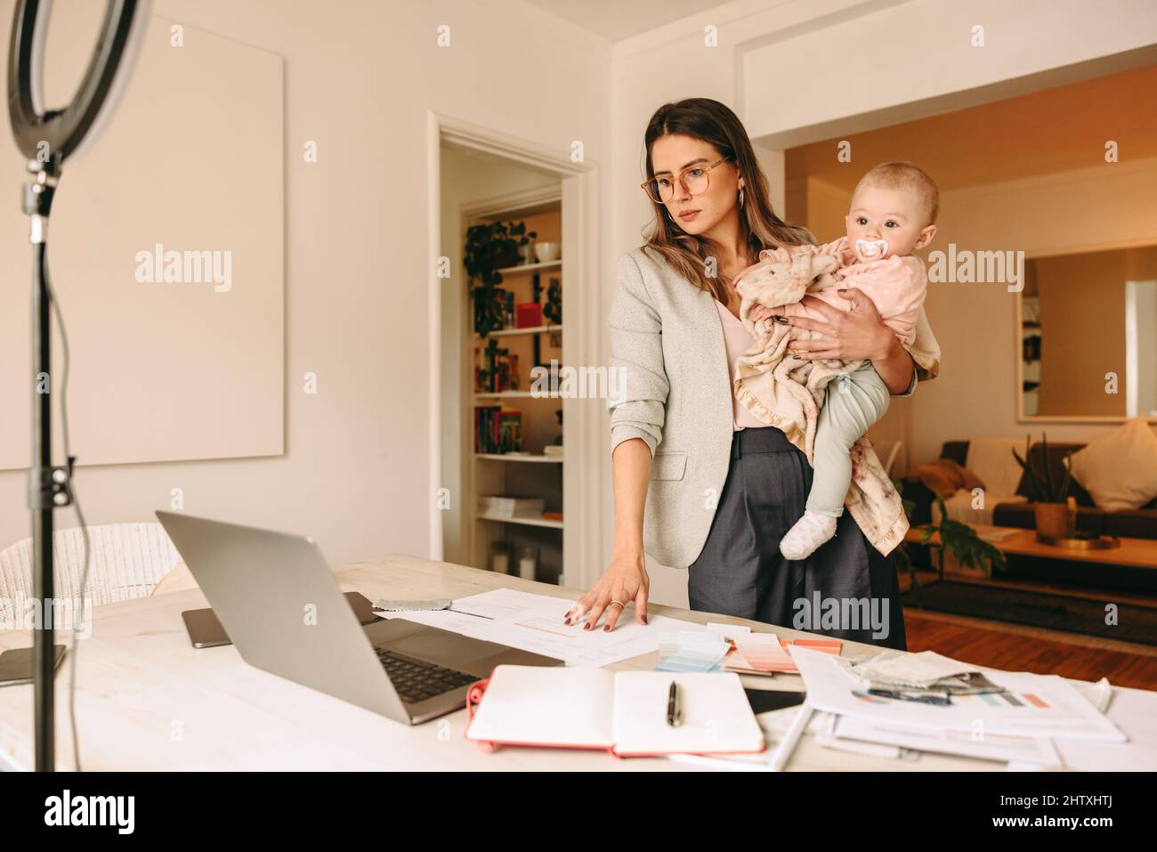 Interior designer holding her baby while working in her home office. Working mom looking at her laptop screen while planning a new project. Female des Stock Photo