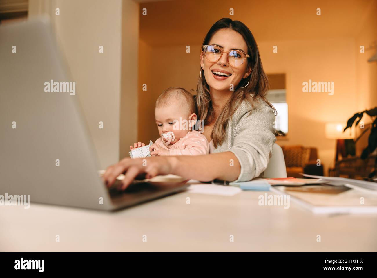 Happy single mom typing on a laptop in her home office. Multitasking mom working on a new creative project at her desk. Female interior designer carry Stock Photo