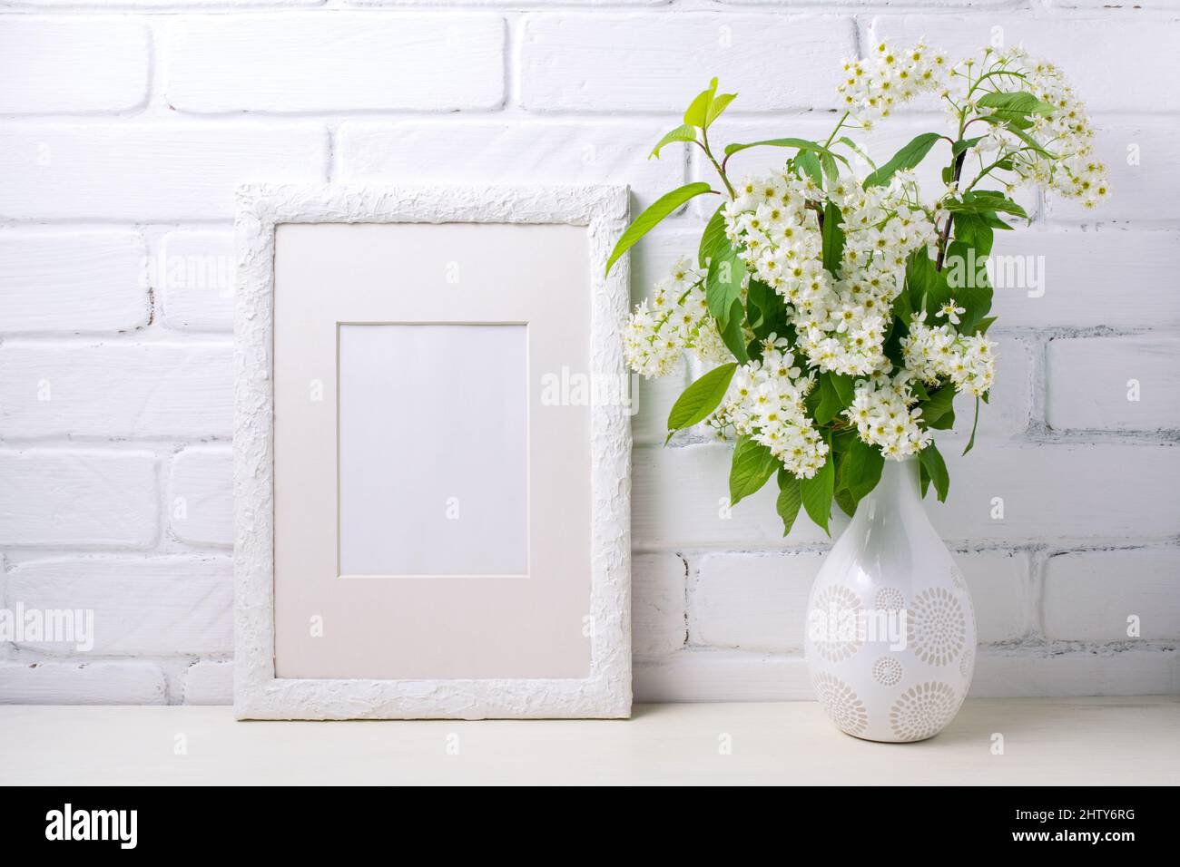 White frame mockup with blooming bird cherry branch near painted brick wall. Empty frame mock up for presentation artwork. Template framing for modern Stock Photo