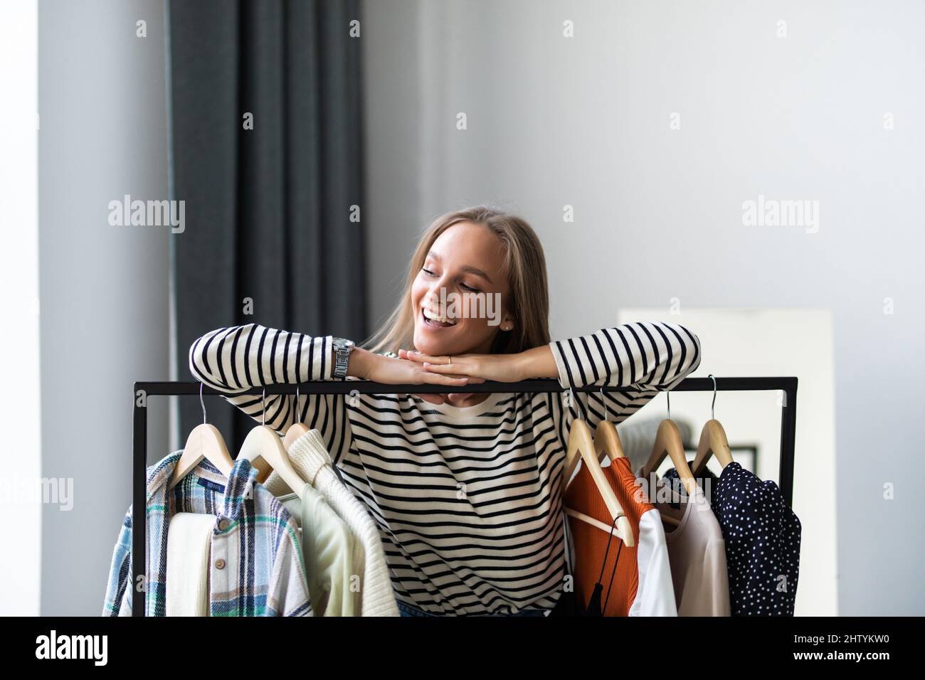 Cute young woman in bathrobe standing in front of hanger rack and trying to choose outfit dressing for work or walk. Selection of a wardrobe, stylist, Stock Photo