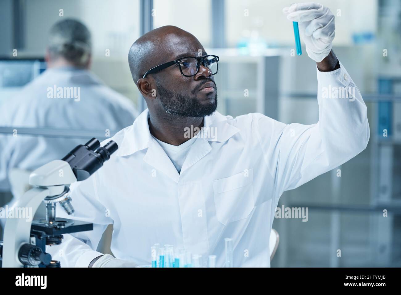 African scientist in eyeglasses and lab coat looking at flask with blue ...