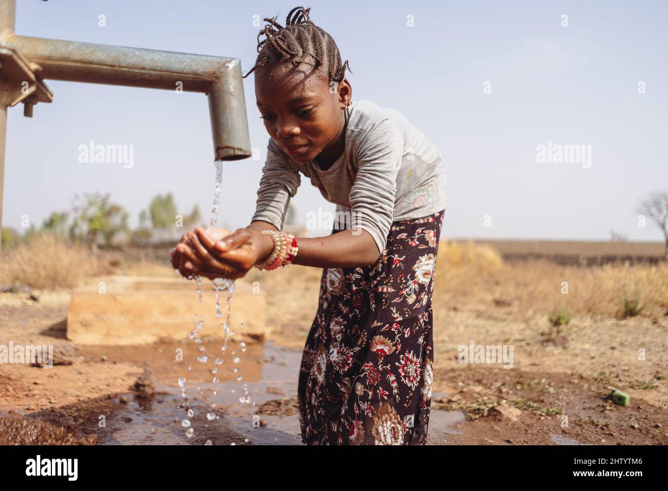 Pretty little black girl hunched over by a West African village pump, happily holding both hands to catch the fresh water running out of the pipe; wat Stock Photo