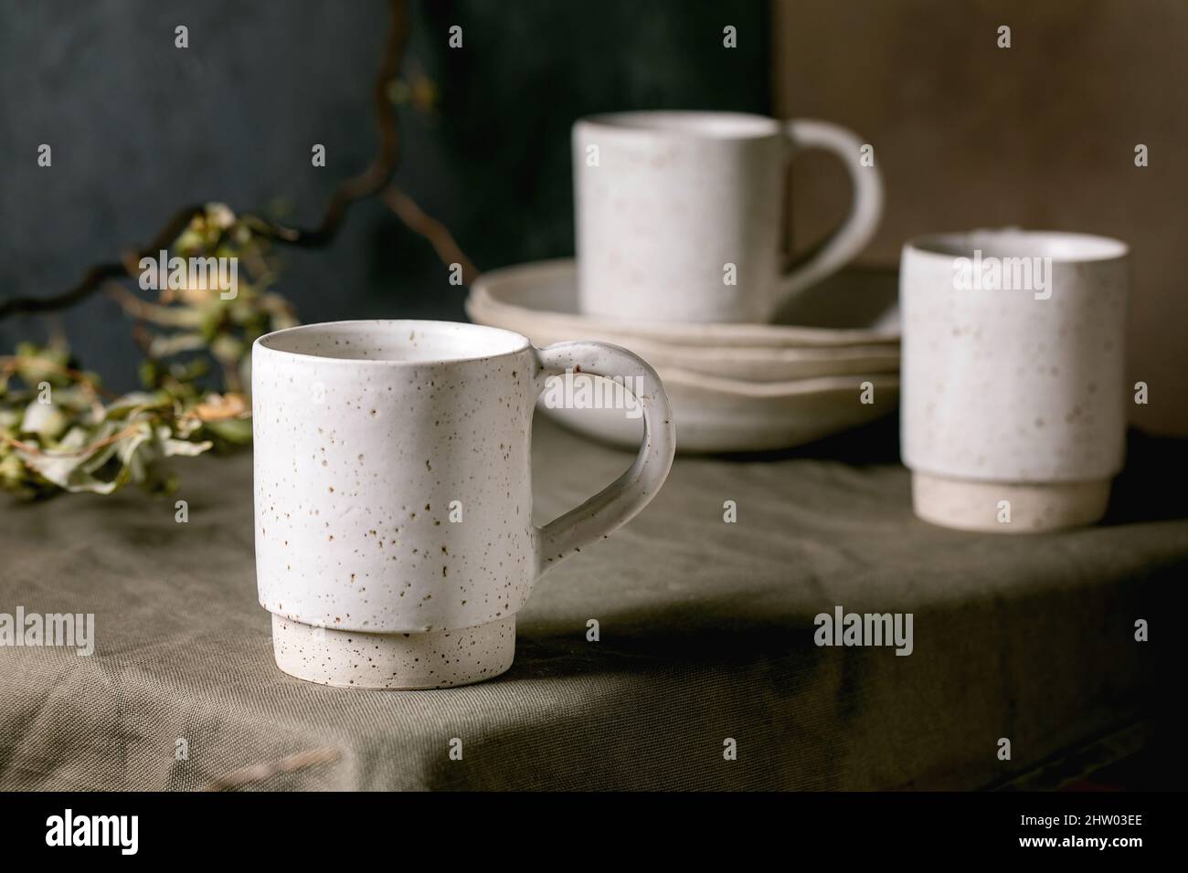 Set of white speckled empty craft ceramic coffee tea mugs standing on dark green linen tablecloth with stack of plates. Stock Photo