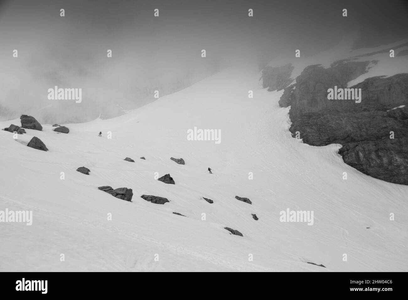 Two tinny skiers in the distance skiing down a slope into the fog Stock Photo