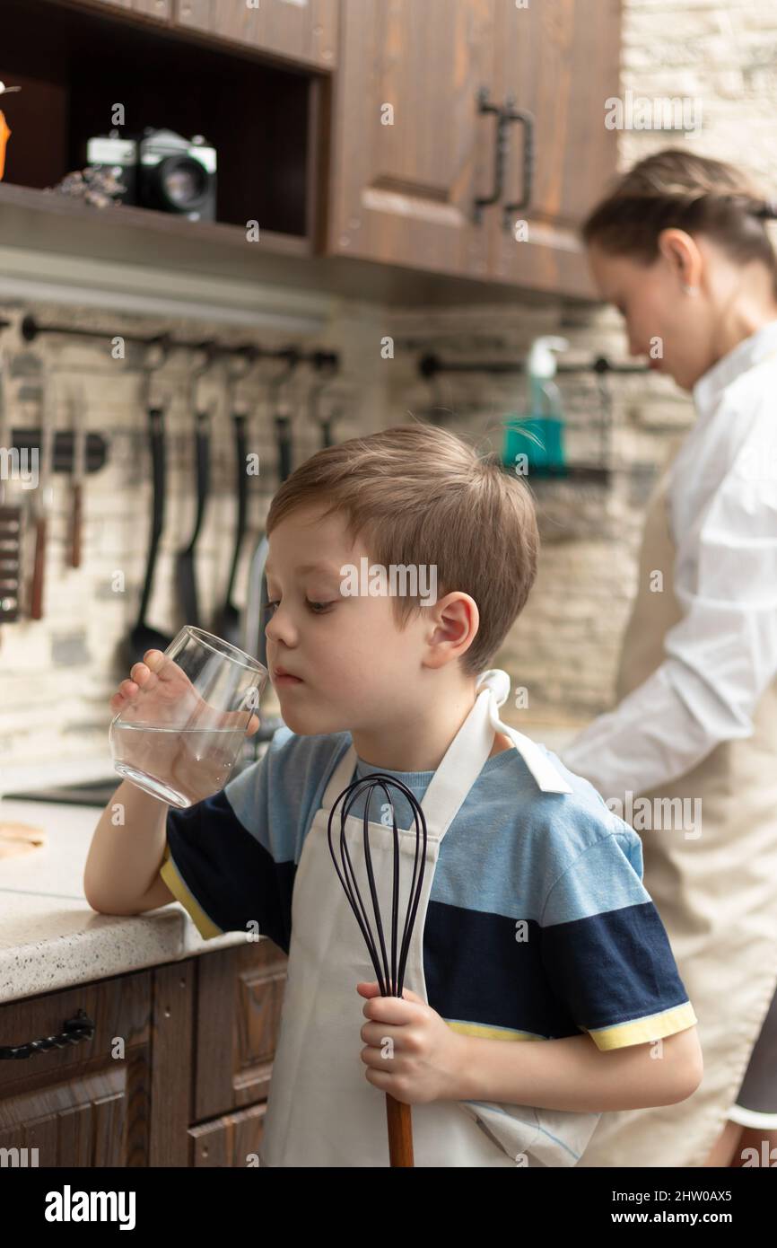 A cute boy of 7 years old in an apron with a whisk in his hand drinks water from a glass in the kitchen against the background of kitchen utensils. Se Stock Photo