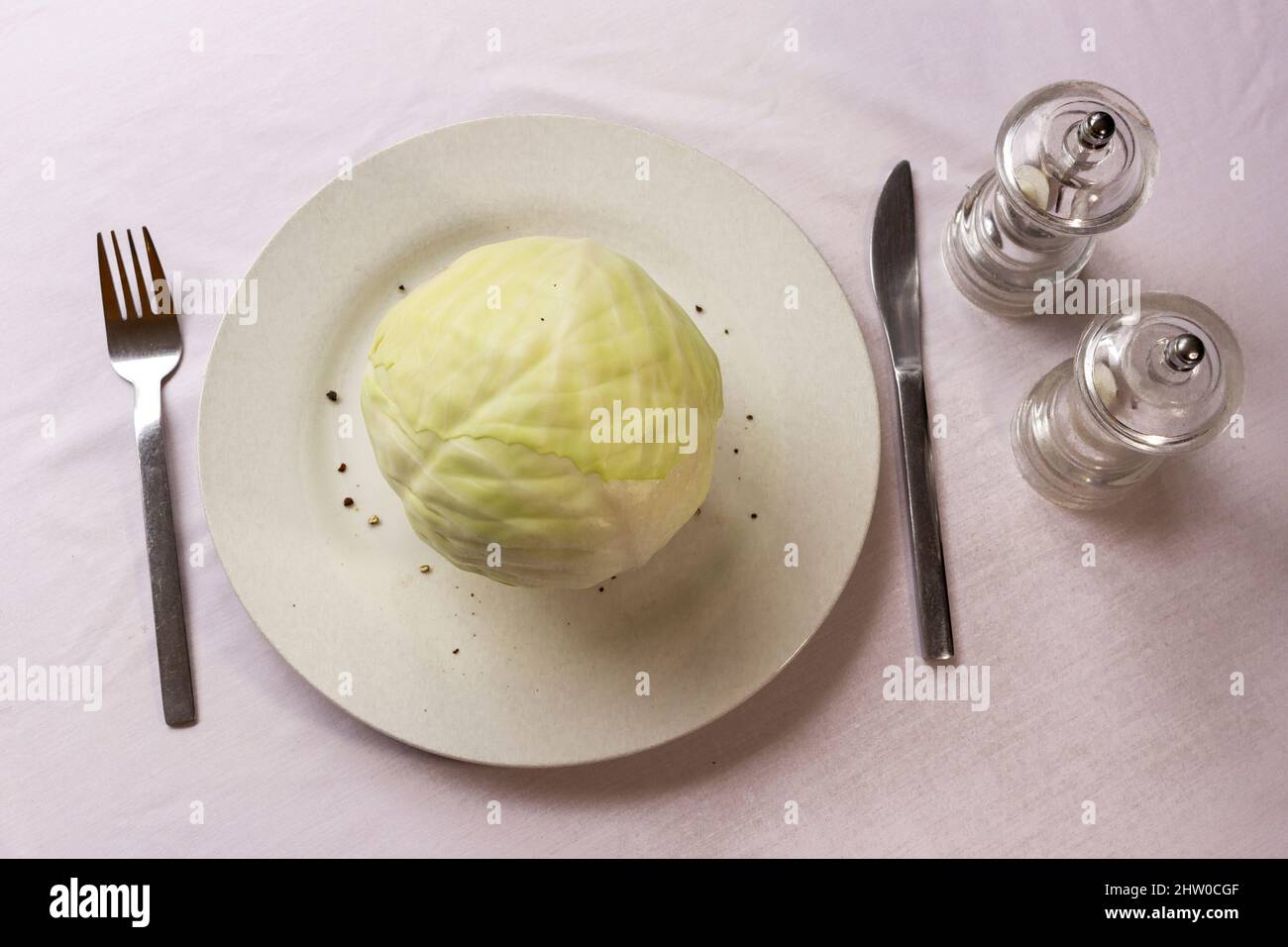 Overhead shot of a a whole white cabbage on white plate with knife and fork and salt and pepper, suggesting a frugal or boring diet Stock Photo