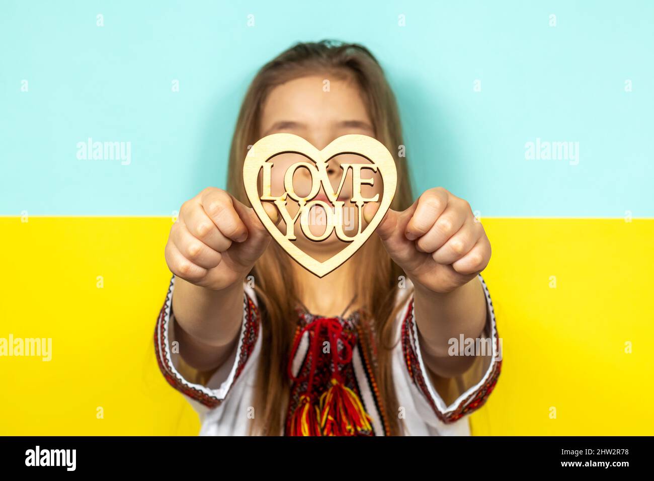 A girl in national Ukrainian clothes, vyshyvanka, holds a wooden heart as a sign of love for Ukraine, close-up against the background of the Ukrainian Stock Photo