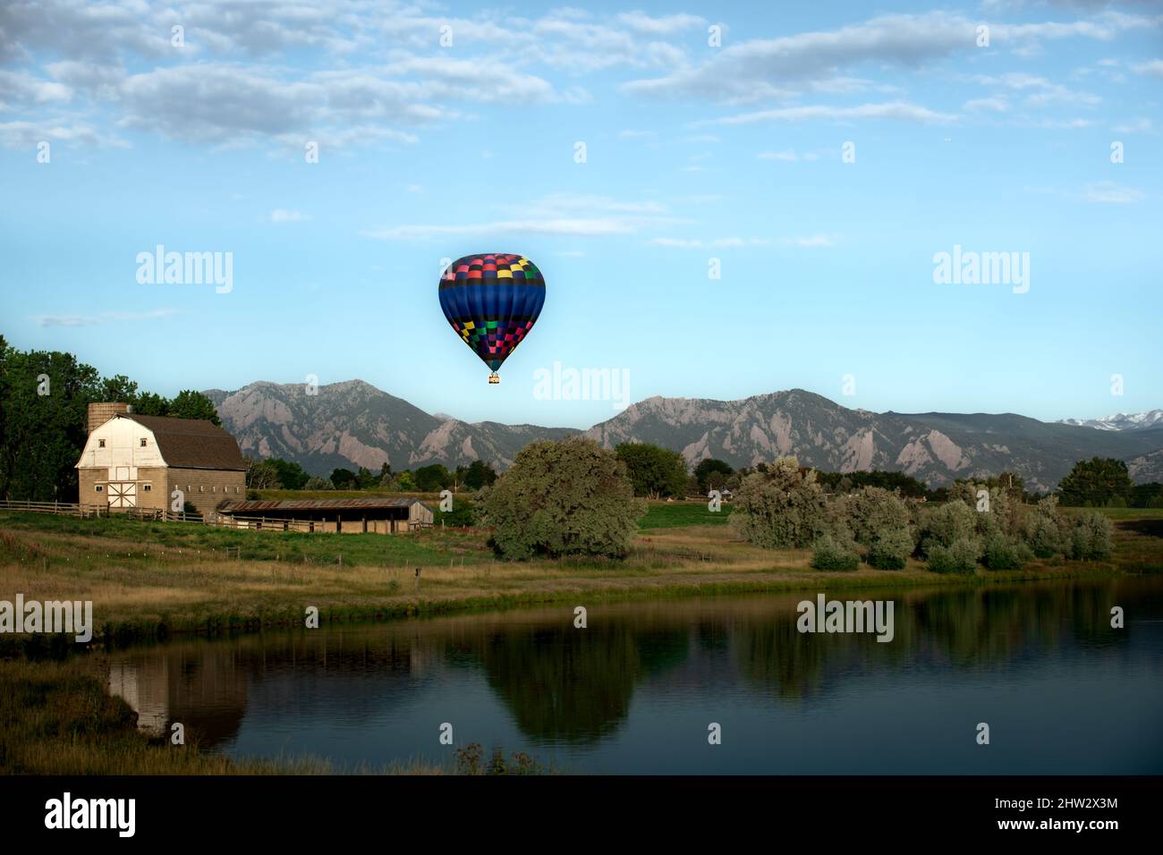 A hot air balloon rises above a barn and a lake with the mountains (the Flatirons) in the background Stock Photo
