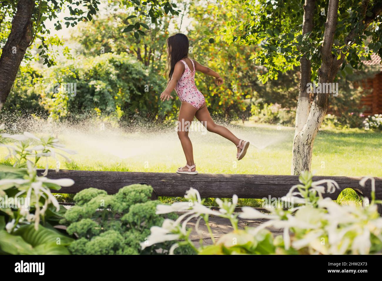 Female child having fun on log finding balance on one foot indulging with water sprinklers, house and green trees in background in daytime. Dreaming Stock Photo