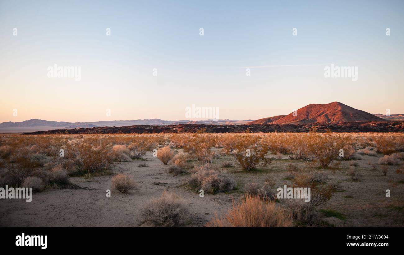 The sun sets on a hill in the distance with a foreground of sage brush in the Mojave Desert Stock Photo