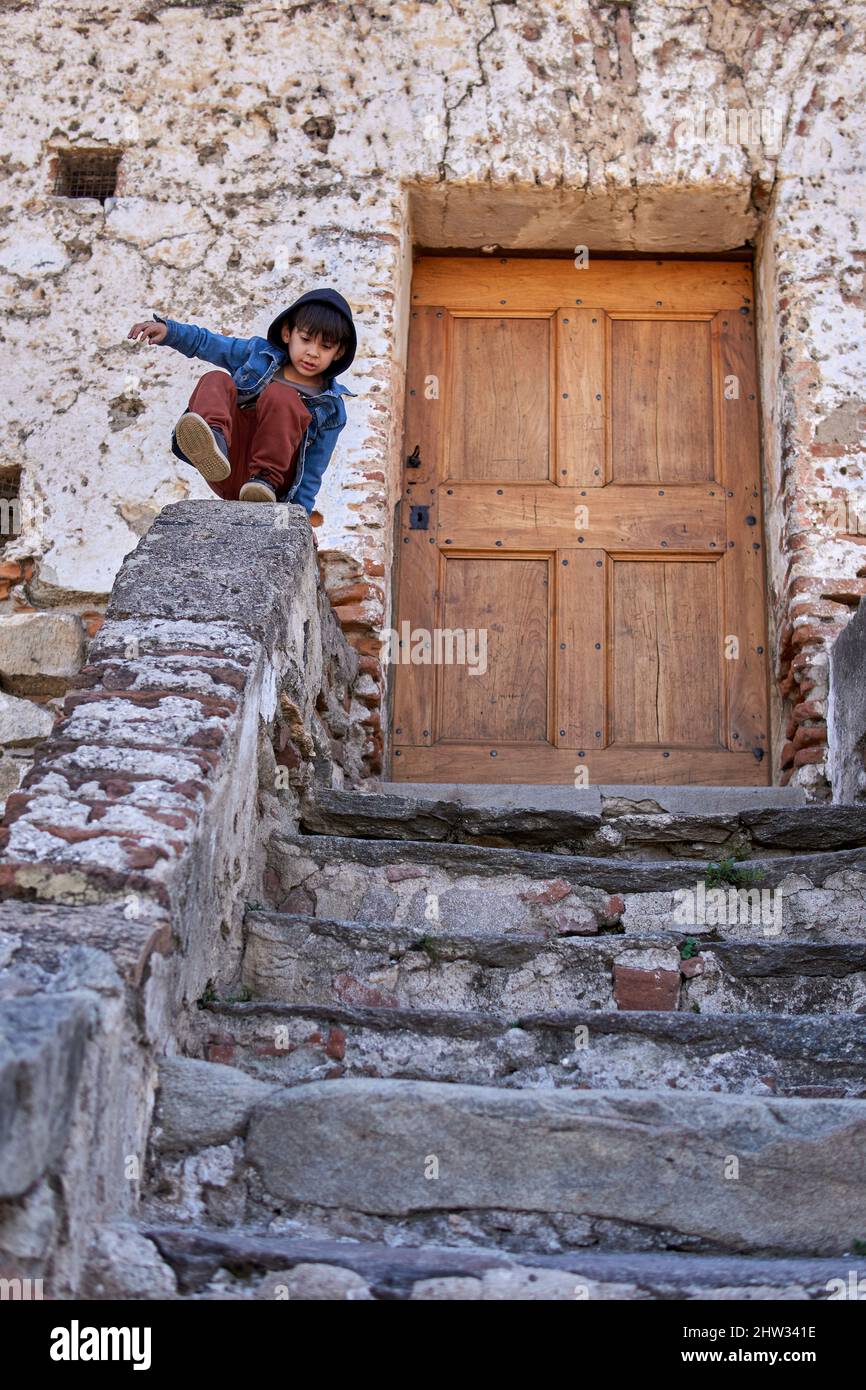 latin kid in casual dress wearing a jean hooded jacket practicing parkour. little boy doing flips on the steps in an old town in Argentina. vintage wo Stock Photo