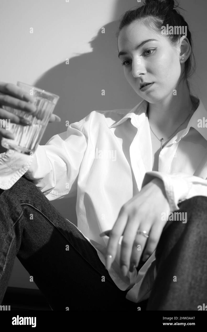 Young woman drinking alcohol from glass against sunny wall background, addiction. monochrome image Stock Photo