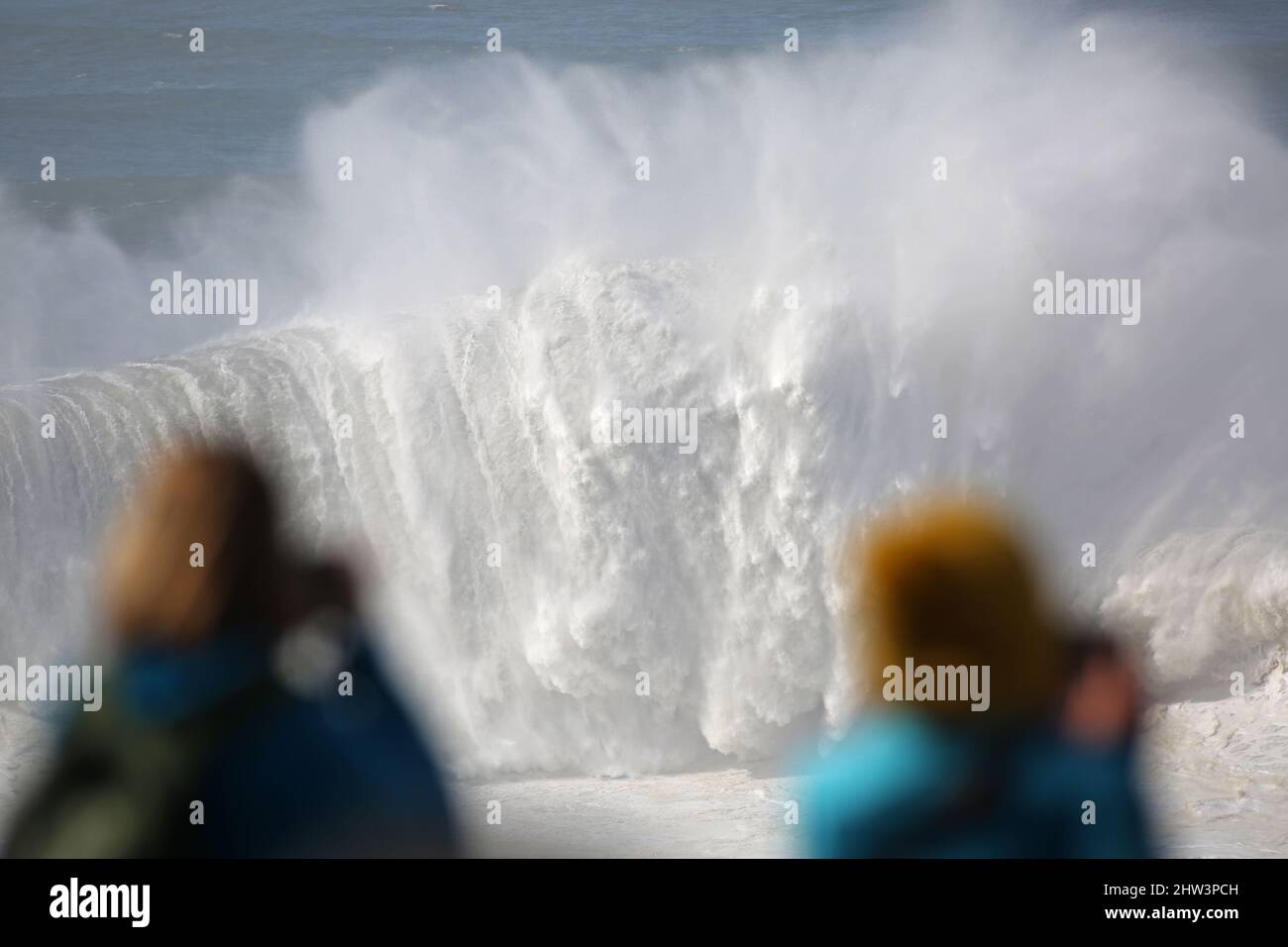 A giant wave breaking while two female spectactors watch and photograph in awe. Praia do Norte, Nazaré, Portugal. Stock Photo