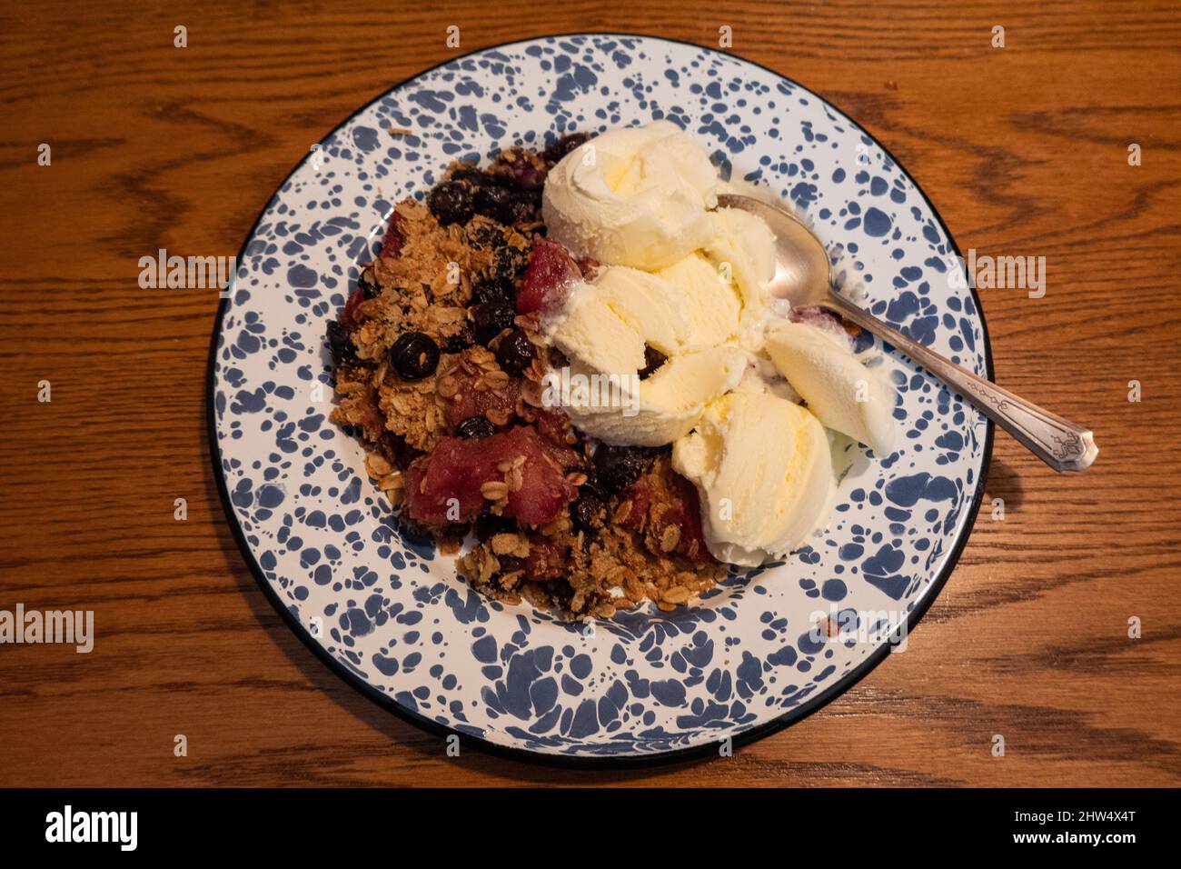 A large serving of apple and blueberry crisp with vanilla ice cream on a blue and white metal plate on an oak table. Stock Photo