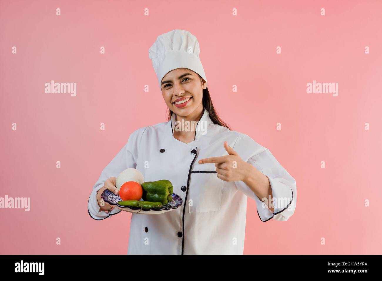 young latin woman wearing chef hat with ingredients on traditional mortar to cook mexican food on pink color background Stock Photo