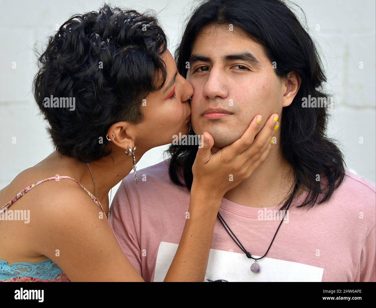 A beautiful Mexican young Latina woman with short hair kisses a handsome Mexican young Latino man with long hair on his cheek. Stock Photo