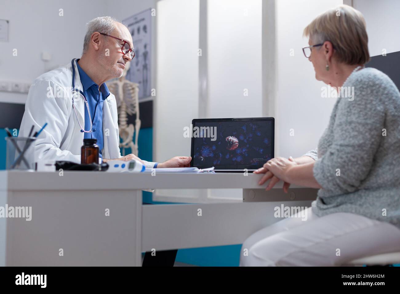 Medic showing coronavirus bacteria illustration on laptop to patient in cabinet. Medical specialist and patient looking at computer screen with visual representation of covid 19 pandemic. Stock Photo