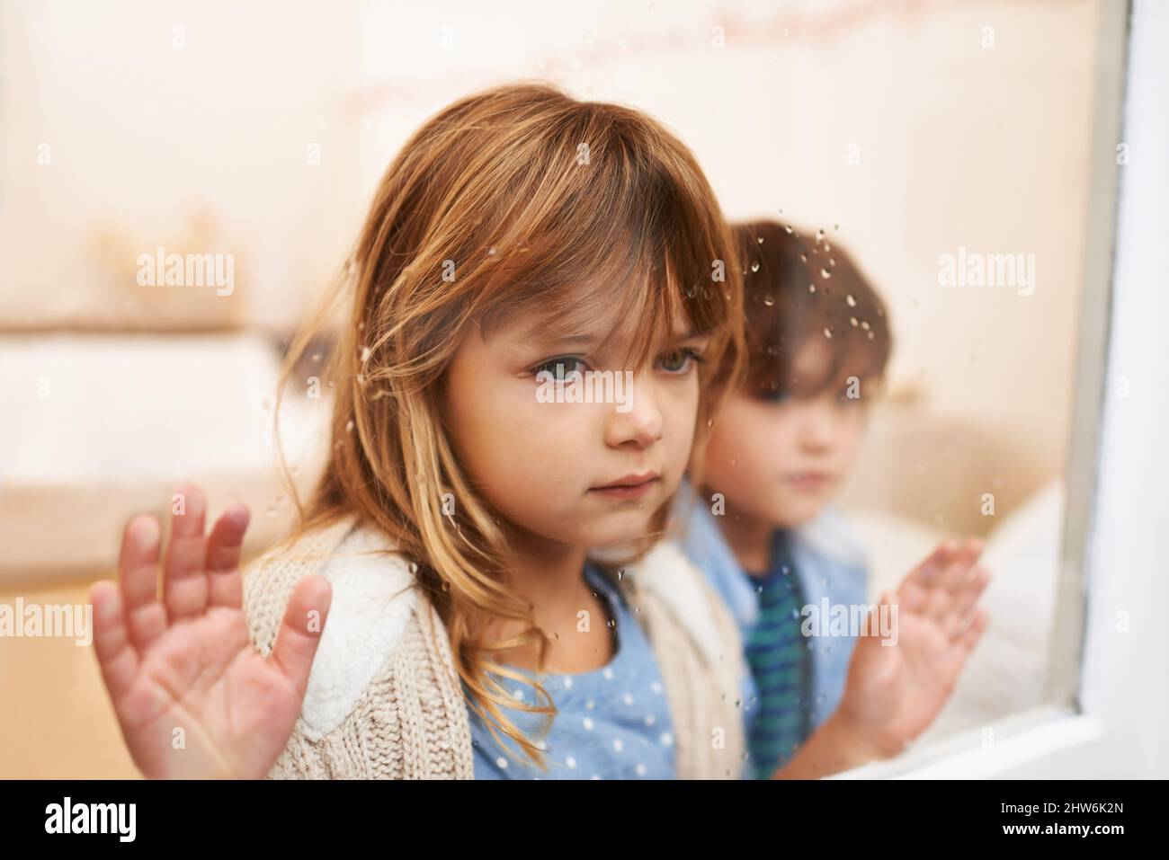 Trapped in a glass box of emotion. Shot of two unhappy-looking young children looking out a window on a rainy day. Stock Photo