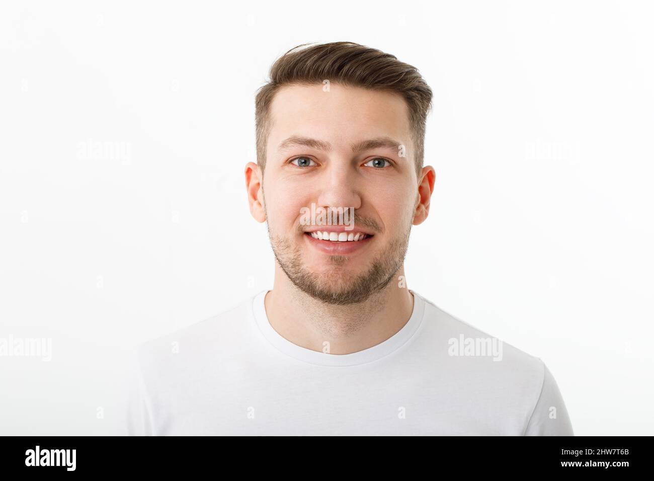 Portrait of a cheerful young man in a white T-shirt on a white background. The guy is standing looking at the camera and smiling. Stock Photo