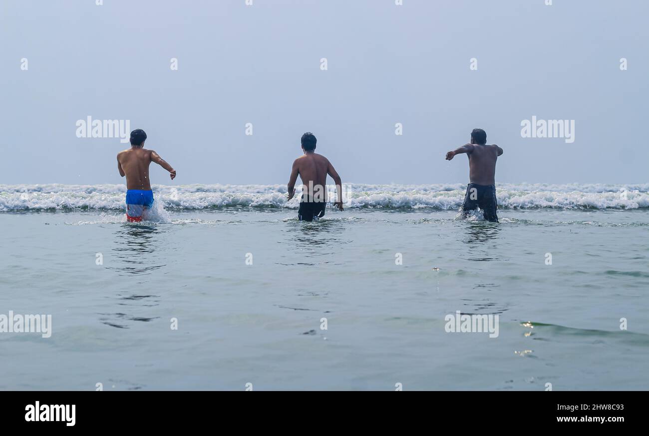 Happy friends are having fun and runs at sunrise beach to sun light and waves. Group of young boys playing together in the ocean waves. Stock Photo