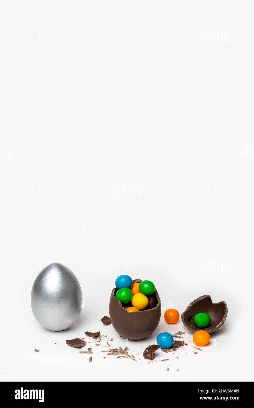 Cracked chocolate easter egg with colorful small round sweets and chocolates and a whole painted silver egg on a white background, copy space, vertica Stock Photo