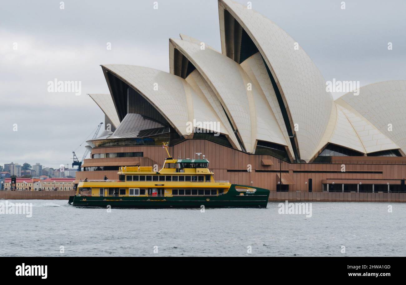 An iconic green and yellow Sydney harbour commuter passenger ferry sails in front of the city's world famous Opera House on a cloudy day Stock Photo