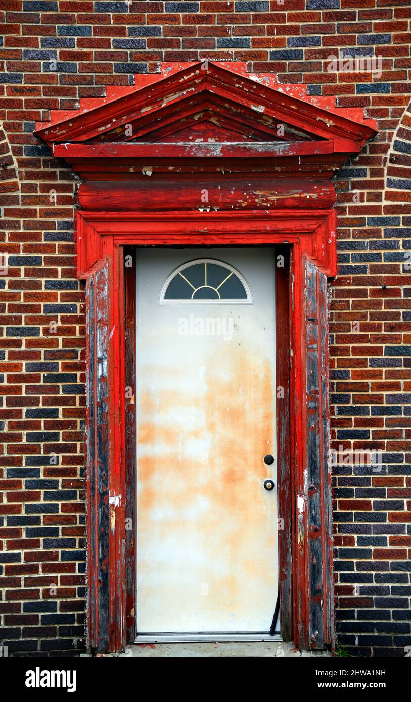Weathered, worn and dilapidated doorway has broken door frame and peeling red paint.  Building is black and red brick. Stock Photo